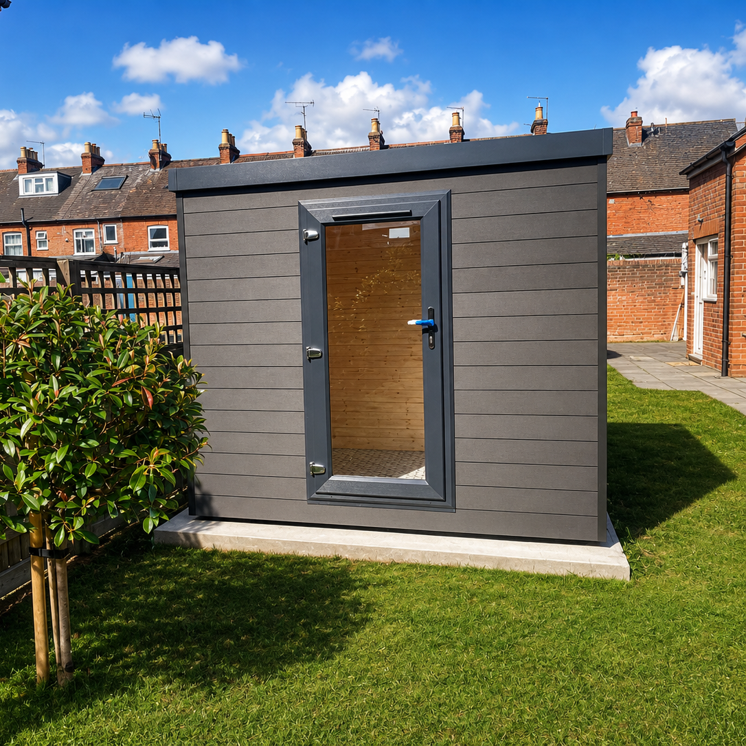A modern gray outdoor garden room with a glass-paneled door, situated on a green lawn in a backyard with brick houses and a blue sky in the background.